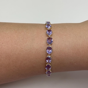 Könnte beinhalten: Ein goldenes Armband mit ovalen, lila Edelsteinen. Die Edelsteine sind in einer Reihe angeordnet und bilden ein zartes Design. Das Armband wird an einem Arm gezeigt.