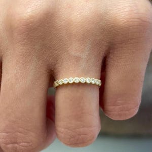Könnte beinhalten: Ein goldfarbener Ring mit einer durchgehenden Reihe kleiner, runder, klarer Edelsteine. Der Ring, der an einem Finger getragen wird, hat eng aneinander liegende Edelsteine, die einen funkelnden Effekt erzeugen. Das Design ist schlicht und elegant. Ringdurchmesser ca. 1,5 cm.