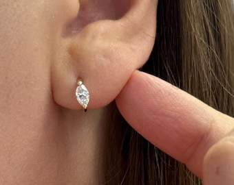 Puede incluir: Un pequeño pendiente de aro dorado con un diamante talla marquesa. El pendiente se lleva en la oreja. El diamante es claro y brillante. El pendiente es de diseño sencillo y elegante.
