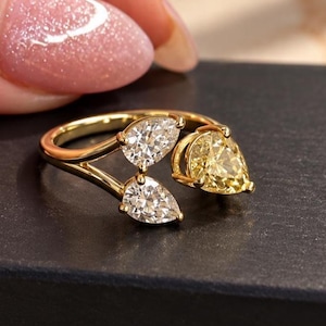Puede incluir: Un anillo de oro con banda abierta que presenta tres piedras de corte en forma de pera. Dos piedras transparentes e incoloras están engastadas en un lado de la banda, mientras que una piedra amarilla se sitúa en el lado opuesto, creando un diseño asimétrico.