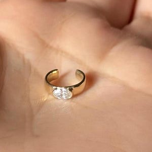 Puede incluir: Un anillo abierto de color dorado con una piedra transparente de forma ovalada en el centro. El anillo está diseñado para ser usado en la oreja o la nariz. El anillo es pequeño y delicado.