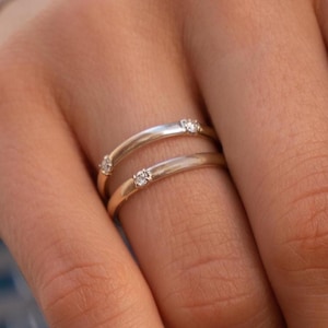 May include: Two silver rings, each with three small, clear gemstones. The rings are stacked on a finger, showcasing their simple yet elegant design. The bands have a polished finish, reflecting light.