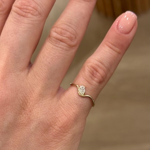 May include: A thin gold ring featuring a single small oval-shaped white gemstone set at the centre of a gentle wave-like curve in the band. The metal band has a smooth, polished finish and follows a delicate, undulating design across the finger.