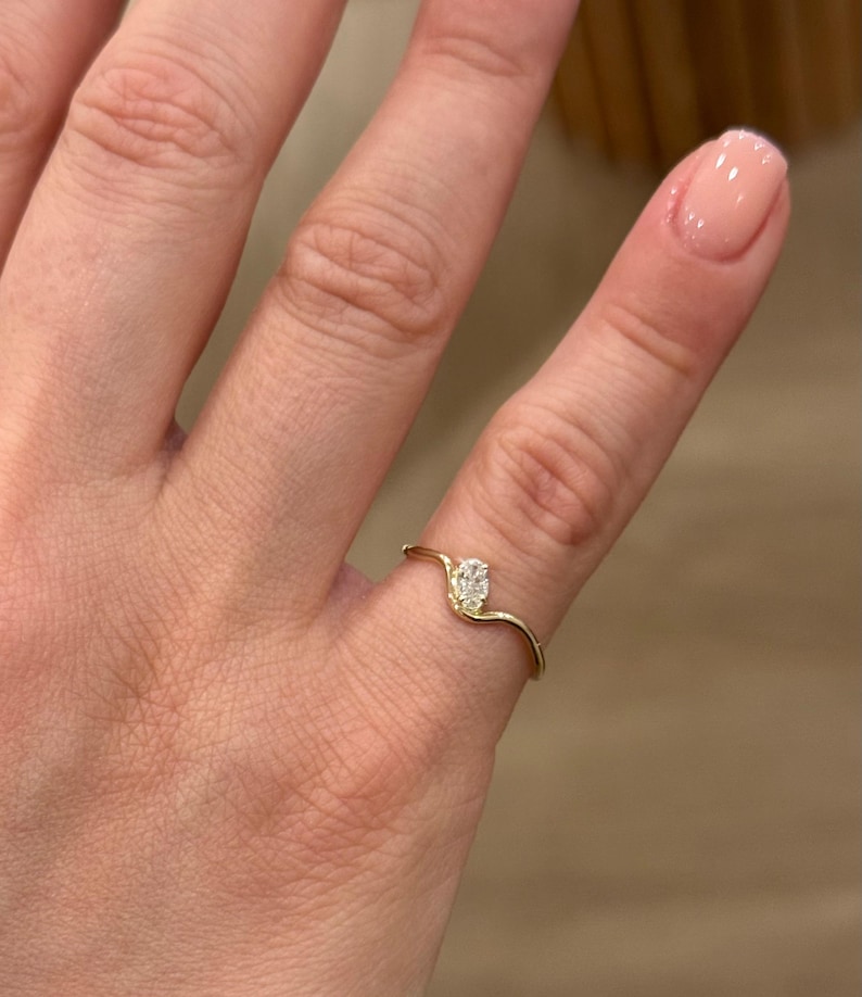 May include: A thin gold ring featuring a single small oval-shaped white gemstone set at the centre of a gentle wave-like curve in the band. The metal band has a smooth, polished finish and follows a delicate, undulating design across the finger.