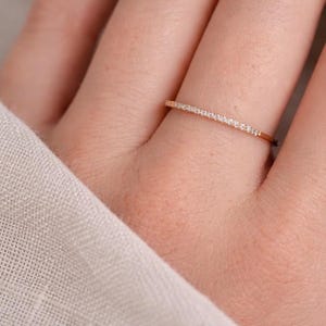 May include: A delicate rose gold ring with a thin band, set with a row of small, sparkling clear gemstones. The ring is shown on a person's finger, with a soft, neutral-colored fabric in the background.