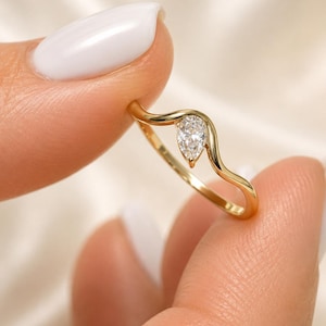 May include: A gold ring featuring a single pear-shaped clear gemstone set within a smooth, wavy band. The metal has a polished yellow finish, and the stone is positioned at the peak of the curved design, creating a delicate and continuous silhouette.