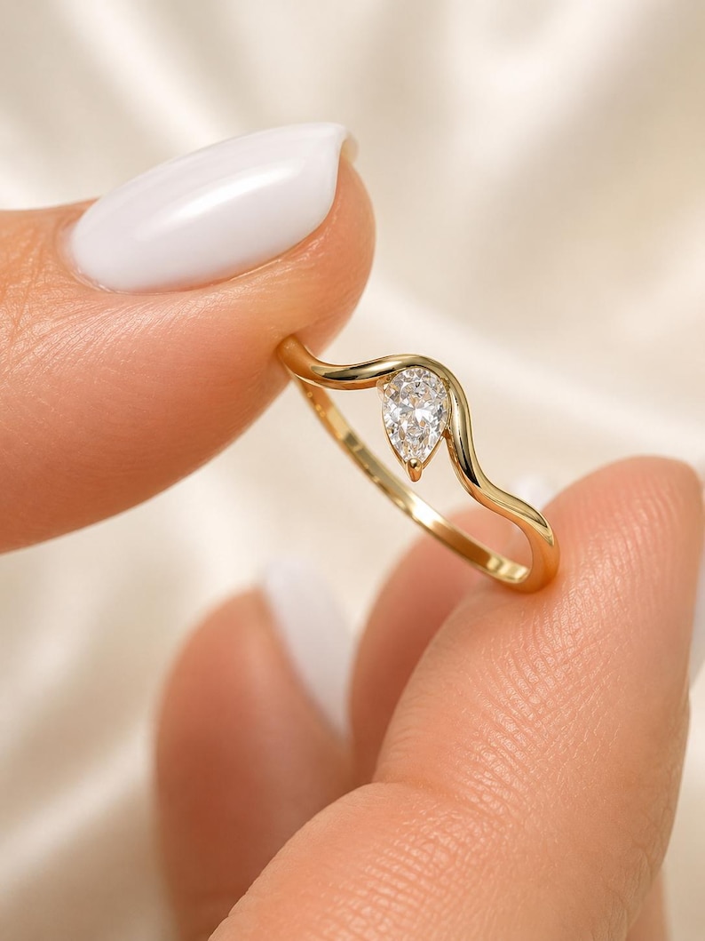 May include: A gold ring featuring a single pear-shaped clear gemstone set within a smooth, wavy band. The metal has a polished yellow finish, and the stone is positioned at the peak of the curved design, creating a delicate and continuous silhouette.