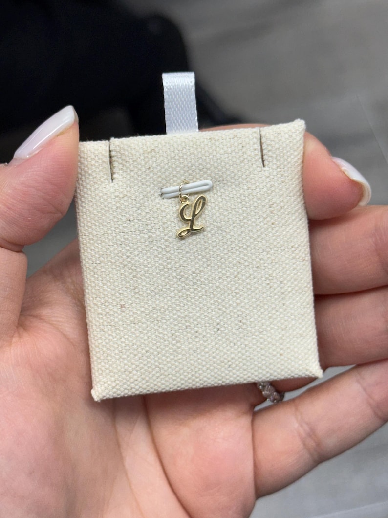 Op de afbeelding: Een goudkleurige hanger in de vorm van de letter "L" wordt getoond op een kleine, cr&egrave;mekleurige stoffen kaart. De hanger is bevestigd aan een kleine staaf met een wit lint. De kaart wordt in een hand gehouden.
