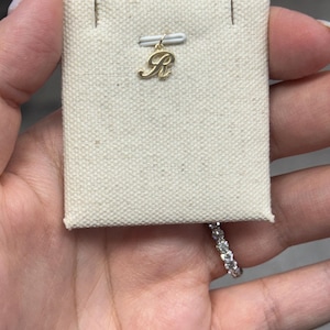 Op de afbeelding: Een goudkleurige hanger in de vorm van de letter "R" wordt getoond op een beige stoffen kaart. De hanger is bevestigd aan een klein wit lint. Een diamanten ring is zichtbaar op de achtergrond.