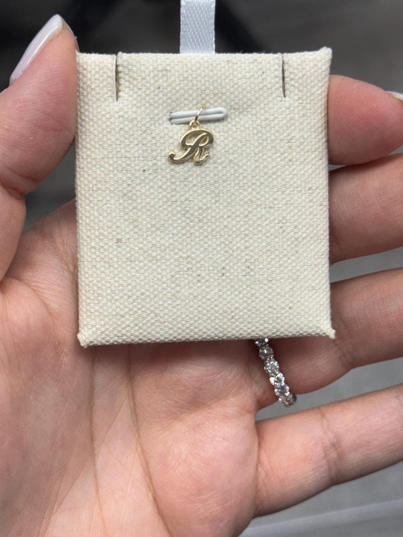 Op de afbeelding: Een goudkleurige hanger in de vorm van de letter "R" wordt getoond op een beige stoffen kaart. De hanger is bevestigd aan een klein wit lint. Een diamanten ring is zichtbaar op de achtergrond.