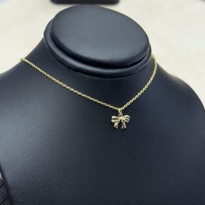 May include: A gold-colored necklace with a bow-shaped pendant. The delicate chain and bow are displayed on a black jewelry stand. The bow pendant is small and detailed.