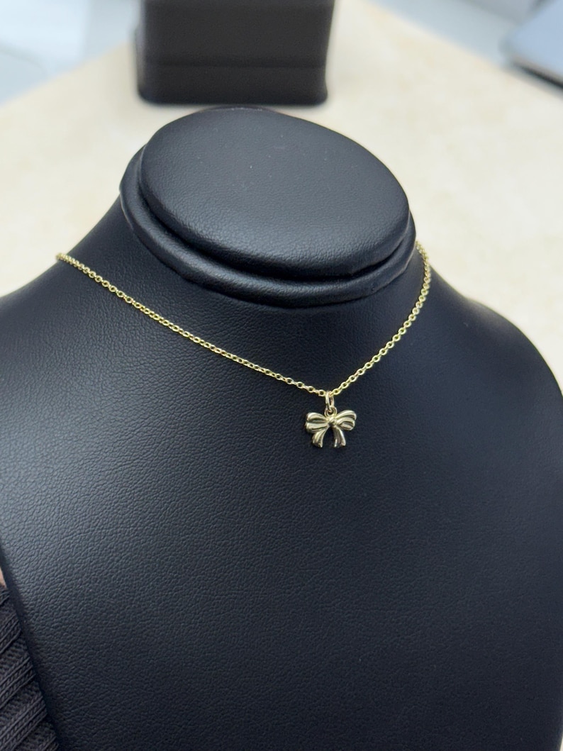 May include: A gold-colored necklace with a bow-shaped pendant. The delicate chain and bow are displayed on a black jewelry stand. The bow pendant is small and detailed.