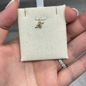 Op de afbeelding: Een goudkleurige letter "K" bedel hanger op een beige stoffen kaart met een wit lint. De bedel is klein en delicaat, met een cursief lettertype. De kaart wordt in een hand gehouden, met een ring zichtbaar op een vinger.