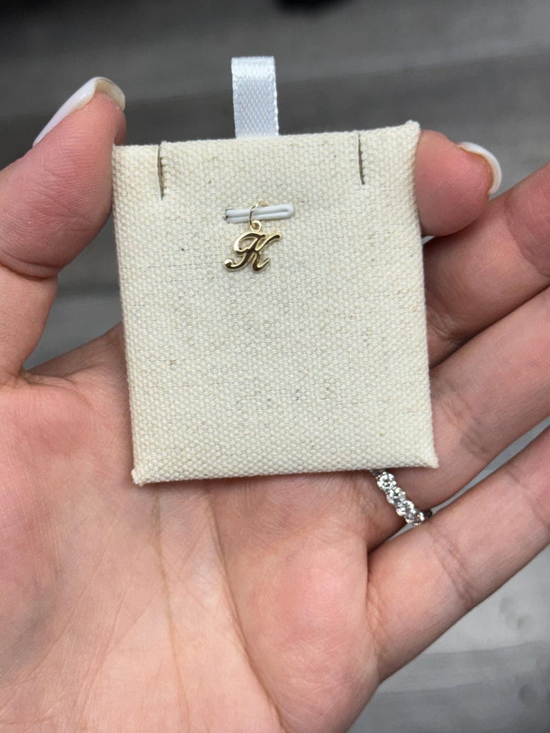 Op de afbeelding: Een goudkleurige letter "K" bedel hanger op een beige stoffen kaart met een wit lint. De bedel is klein en delicaat, met een cursief lettertype. De kaart wordt in een hand gehouden, met een ring zichtbaar op een vinger.
