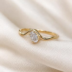May include: A gold ring featuring a single oval-cut clear gemstone set in a wavy, curved band. The metal has a polished finish, and the stone is held in place by four small prongs, creating a continuous, flowing design along the finger band.