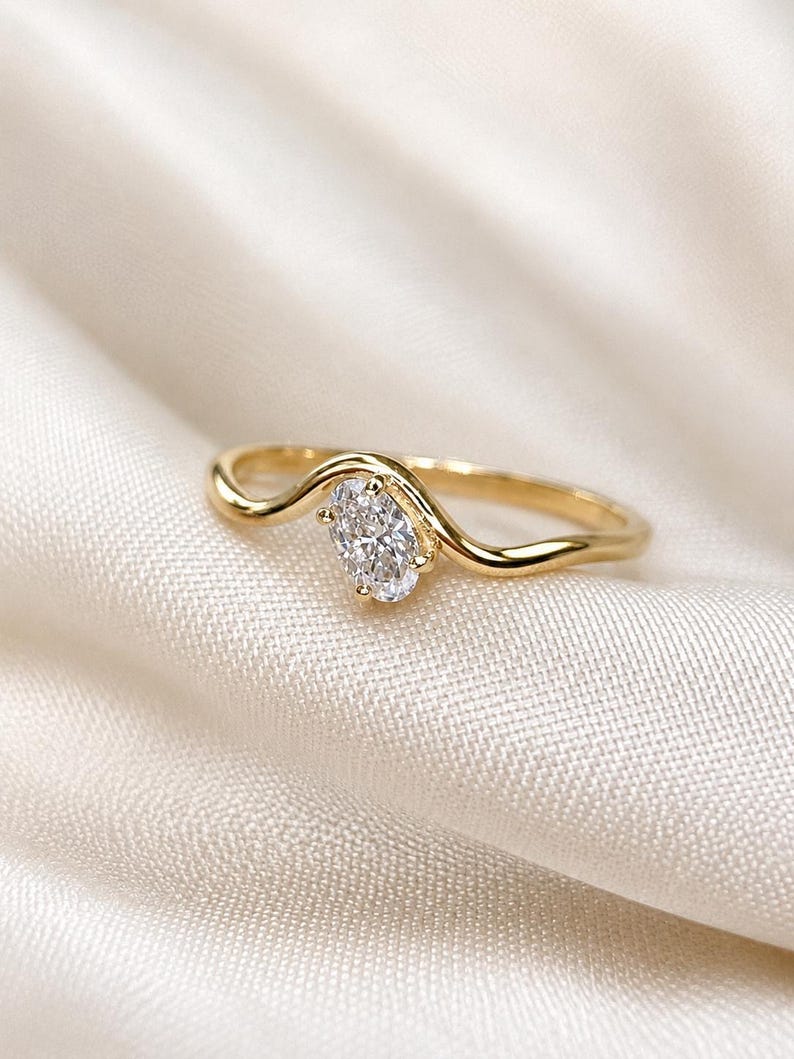 May include: A gold ring featuring a single oval-cut clear gemstone set in a wavy, curved band. The metal has a polished finish, and the stone is held in place by four small prongs, creating a continuous, flowing design along the finger band.