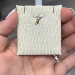 Op de afbeelding: Een kleine, goudkleurige hanger in de vorm van de letter "J" wordt getoond op een cr&egrave;mekleurige stoffen kaart. De hanger is bevestigd aan een kleine goudkleurige sluiting. De stoffen kaart wordt door een hand vastgehouden.