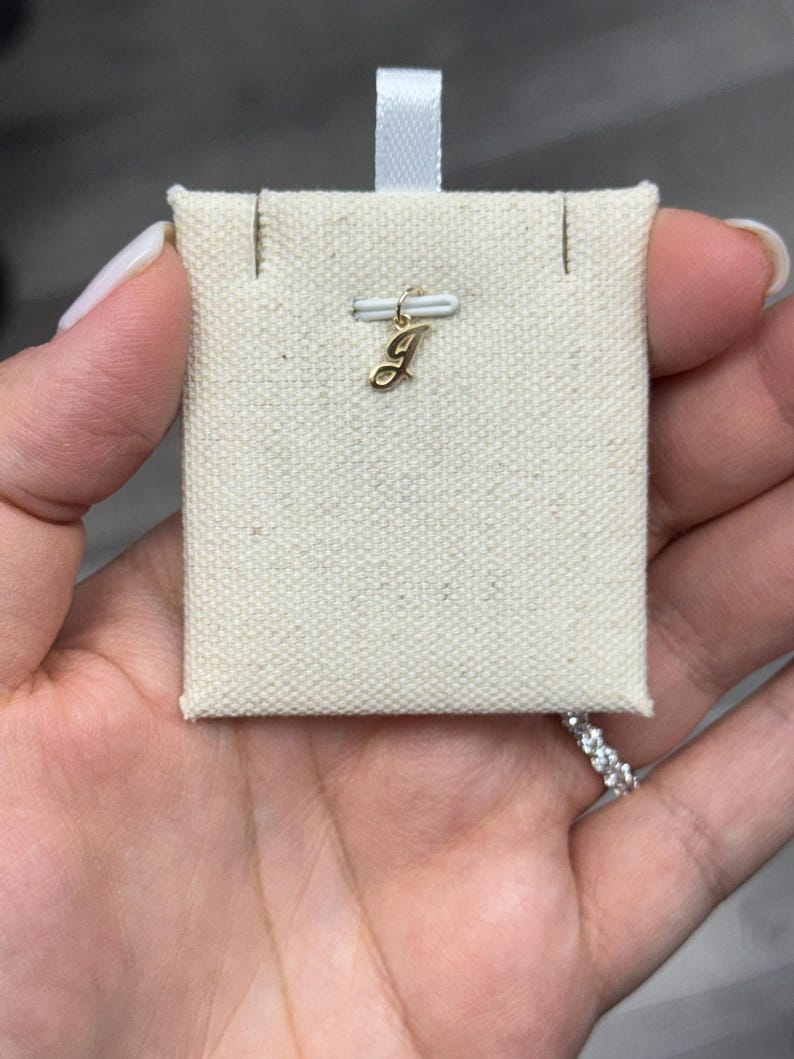 Op de afbeelding: Een kleine, goudkleurige hanger in de vorm van de letter "J" wordt getoond op een cr&egrave;mekleurige stoffen kaart. De hanger is bevestigd aan een kleine goudkleurige sluiting. De stoffen kaart wordt door een hand vastgehouden.