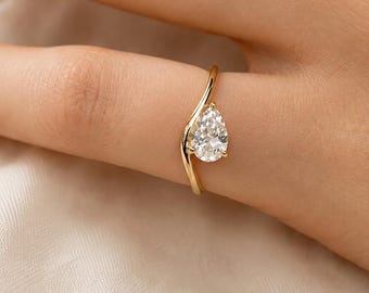 May include: A gold ring featuring a single pear-shaped clear gemstone set on a slender, curved band. The metal band has a smooth, polished finish and follows a gentle wave-like contour that leads to the faceted stone held by small prongs.