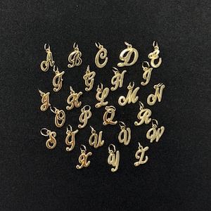 May include: A selection of gold-coloured cursive alphabet letter charms, each with a small loop at the top for attaching to a chain or bracelet. The letters are displayed on a dark background.