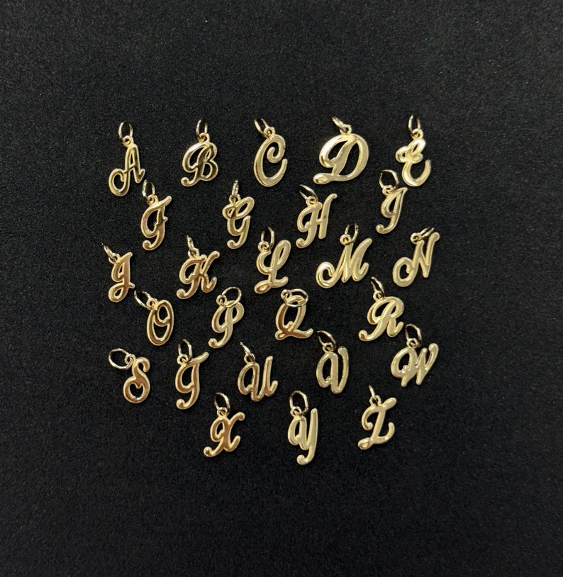 Op de afbeelding: Een verzameling goudkleurige alfabetletters in sierlijke letters, elk met een kleine lus aan de bovenkant om aan een ketting of armband te bevestigen. De letters worden op een donkere achtergrond weergegeven.