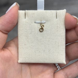 Op de afbeelding: Een goudkleurige initiaalbedel in de vorm van de letter "S" wordt getoond op een cr&egrave;mekleurige stoffen kaart. De bedel is bevestigd aan een kleine witte staaf. De kaart heeft een wit lint aan de bovenkant.