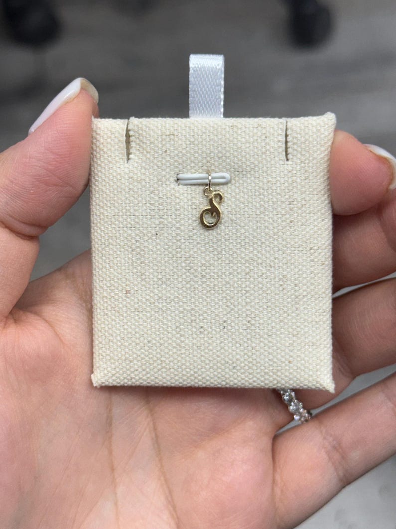 Op de afbeelding: Een goudkleurige initiaalbedel in de vorm van de letter "S" wordt getoond op een cr&egrave;mekleurige stoffen kaart. De bedel is bevestigd aan een kleine witte staaf. De kaart heeft een wit lint aan de bovenkant.