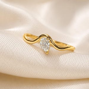 May include: A gold ring featuring a single pear-shaped clear gemstone set in a curved, wave-like band. The metal has a polished, reflective finish, and the stone is positioned at the centre of the band's peak, showcasing a faceted surface.