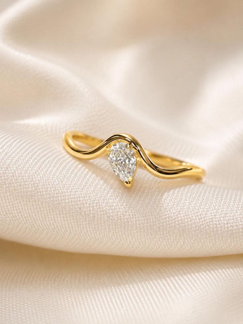 May include: A gold ring featuring a single pear-shaped clear gemstone set in a curved, wave-like band. The metal has a polished, reflective finish, and the stone is positioned at the centre of the band's peak, showcasing a faceted surface.