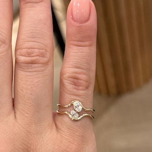 May include: A gold ring featuring two wavy, parallel bands that meet in the centre. The ring is set with two small, clear, pear-shaped stones positioned vertically between the two bands, creating an open, bypass-style design on the finger.