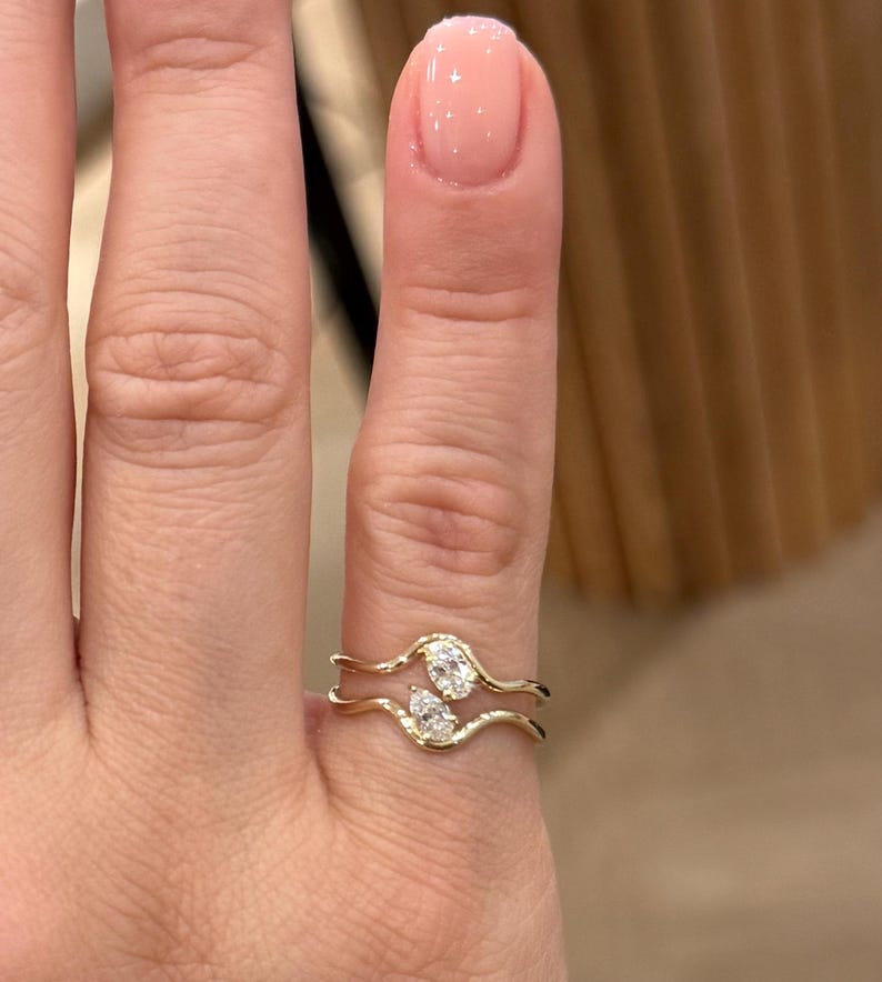 May include: A gold ring featuring two wavy, parallel bands that meet in the centre. The ring is set with two small, clear, pear-shaped stones positioned vertically between the two bands, creating an open, bypass-style design on the finger.