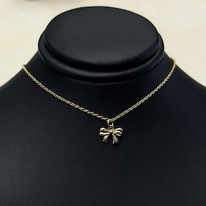 May include: A delicate gold-colored necklace featuring a small bow pendant. The bow is centered on a fine chain, displayed against a black background. The necklace is presented on a black jewelry display.