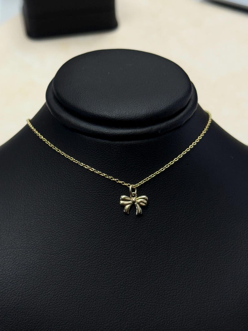 May include: A delicate gold-colored necklace featuring a small bow pendant. The bow is centered on a fine chain, displayed against a black background. The necklace is presented on a black jewelry display.