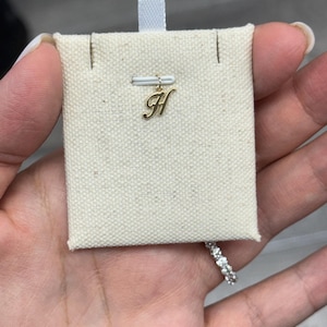 Op de afbeelding: Een goudkleurige letter "H" bedel hanger, opgehangen aan een kleine, off-white stoffen displaykaart. De bedel is bevestigd aan een dunne, goudkleurige staaf. Een diamanten ring is zichtbaar in de rechter benedenhoek.