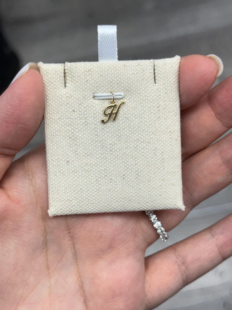 Op de afbeelding: Een goudkleurige letter "H" bedel hanger, opgehangen aan een kleine, off-white stoffen displaykaart. De bedel is bevestigd aan een dunne, goudkleurige staaf. Een diamanten ring is zichtbaar in de rechter benedenhoek.