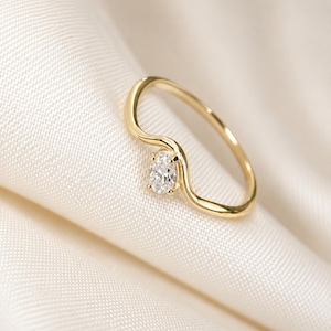 May include: A gold ring featuring a delicate, wavy band design with a single, small, oval-cut clear gemstone set securely at the centre of the curve. The metal has a polished, reflective finish, and the stone is faceted to catch the light.