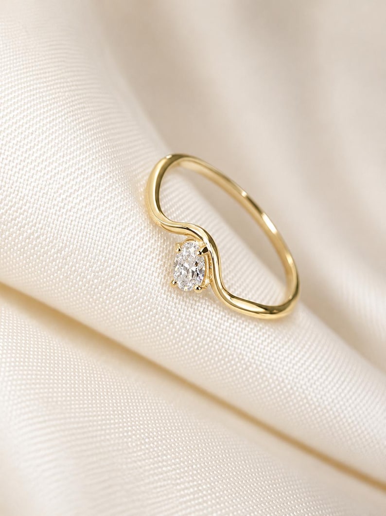 May include: A gold ring featuring a delicate, wavy band design with a single, small, oval-cut clear gemstone set securely at the centre of the curve. The metal has a polished, reflective finish, and the stone is faceted to catch the light.