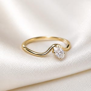 May include: A gold ring featuring a smooth, curved band that dips into a wave-like shape. A single, clear, oval-cut gemstone is set at the centre of the curve, held in place by a small metal prong setting that secures the stone to the band.