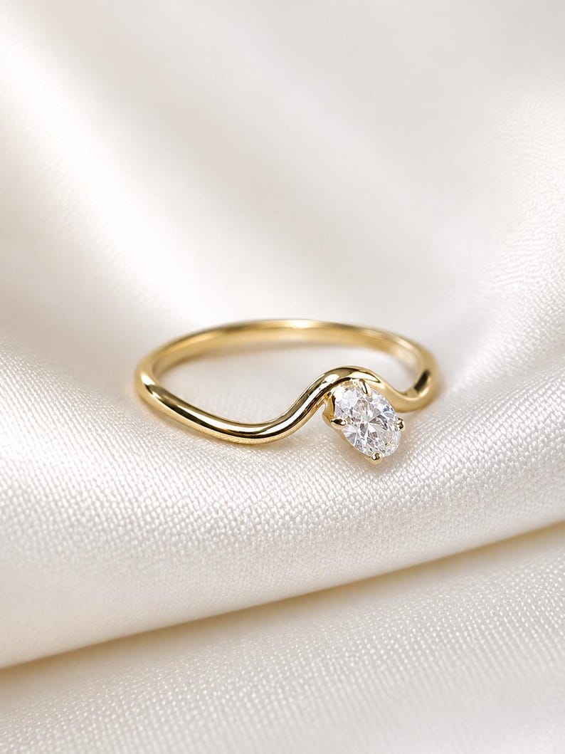 May include: A gold ring featuring a smooth, curved band that dips into a wave-like shape. A single, clear, oval-cut gemstone is set at the centre of the curve, held in place by a small metal prong setting that secures the stone to the band.