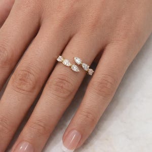 Puede incluir: Un anillo dorado con un diseño de bypass, adornado con múltiples piedras preciosas claras en forma de pera. El anillo se lleva en un dedo, mostrando las piedras brillantes.
