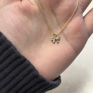 May include: A delicate gold-colored necklace featuring a small bow pendant. The bow is centered on a fine chain, creating a simple and elegant design. The necklace is shown against a neutral background.
