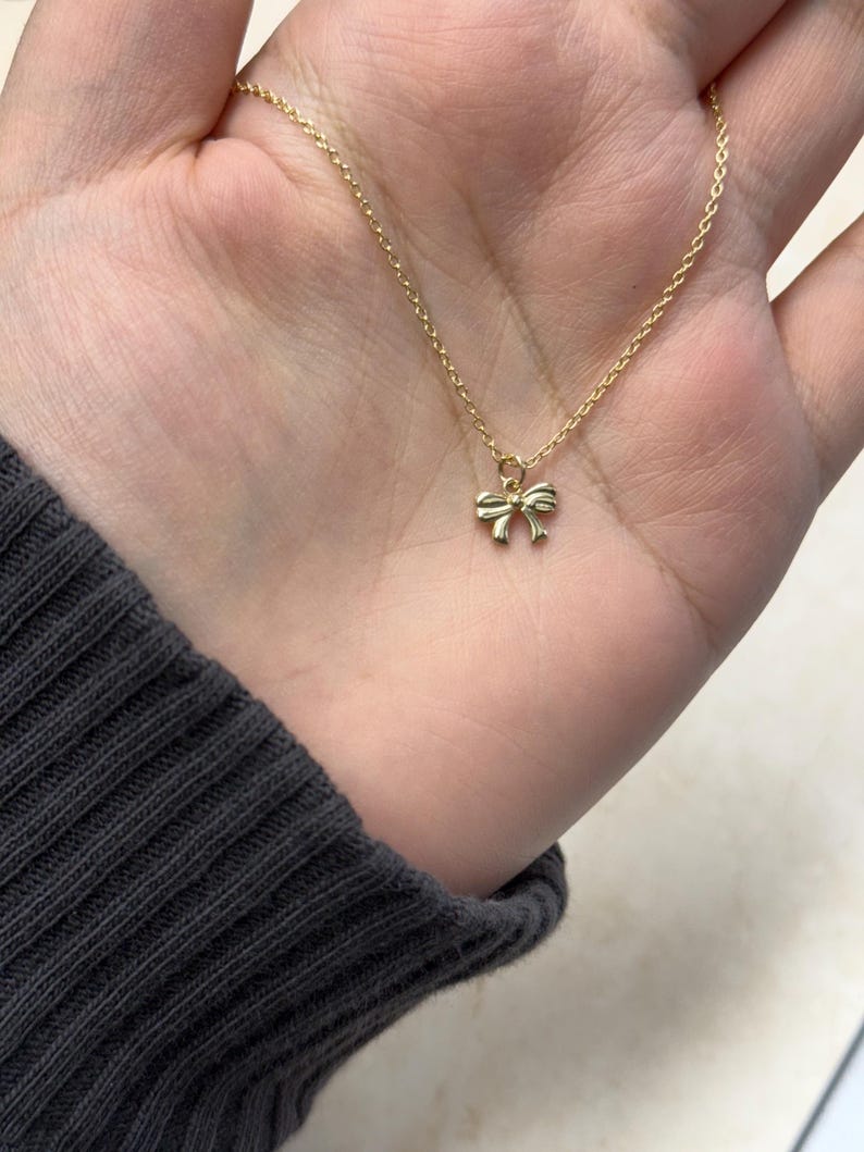 May include: A delicate gold-colored necklace featuring a small bow pendant. The bow is centered on a fine chain, creating a simple and elegant design. The necklace is shown against a neutral background.