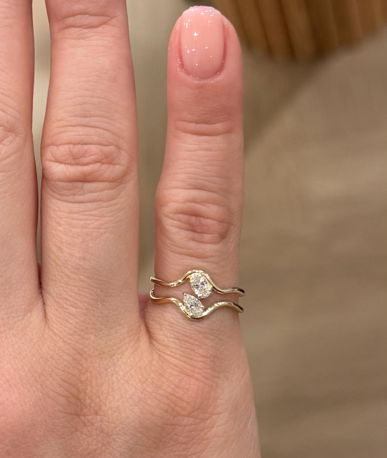May include: A gold bypass ring featuring two distinct bands that curve around the finger. The ring is set with two clear, faceted stones: one oval-shaped stone on the upper band and one pear-shaped stone on the lower band, creating a balanced design.