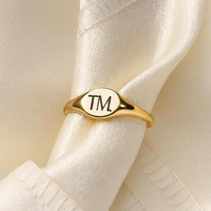 May include: A gold ring featuring a circular face with the letters "TM" in black. The ring is displayed against a white background, highlighting its design.