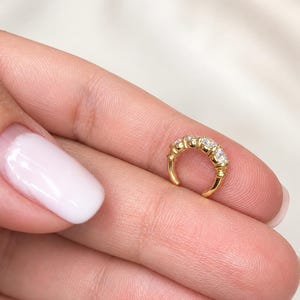 May include: A gold ear cuff featuring a row of five small, clear, round-cut stones set along the outer curve. The metal band has a polished finish, and the design is open, allowing it to be worn on the cartilage of the ear without a piercing.