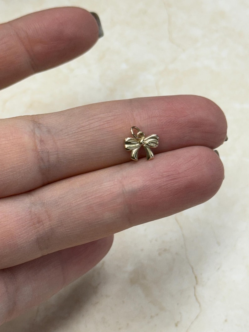 May include: A small, gold-colored bow charm. The charm is intricately designed with detailed curves and a loop at the top for attachment. The bow is a classic shape, suitable for jewelry making or other craft projects.
