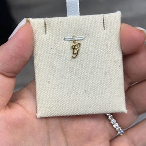 Op de afbeelding: Een goudkleurige bedel in de vorm van de letter "G" wordt getoond op een kleine, cr&egrave;mekleurige stoffen kaart. De bedel is opgehangen aan een kleine metalen lus. Een wit lint is aan de bovenkant van de kaart bevestigd. De bedel wordt in een hand gehouden.