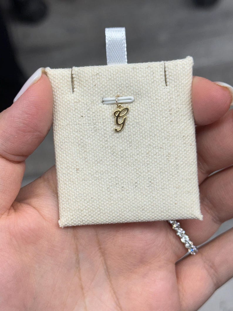 Op de afbeelding: Een goudkleurige bedel in de vorm van de letter "G" wordt getoond op een kleine, cr&egrave;mekleurige stoffen kaart. De bedel is opgehangen aan een kleine metalen lus. Een wit lint is aan de bovenkant van de kaart bevestigd. De bedel wordt in een hand gehouden.