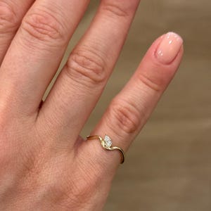 May include: A thin gold ring featuring a delicate, wavy band design that curves upwards to hold a single, small, pear-shaped white gemstone. The metal has a polished, reflective finish, and the stone is set securely at the peak of the band's curve.