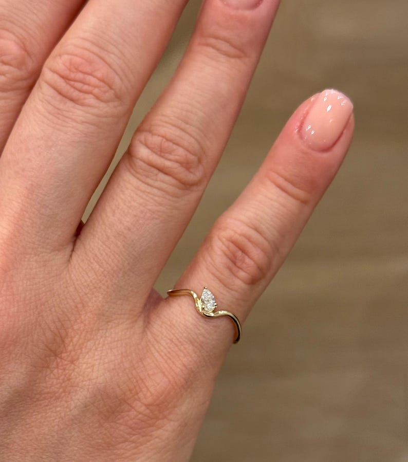 May include: A thin gold ring featuring a delicate, wavy band design that curves upwards to hold a single, small, pear-shaped white gemstone. The metal has a polished, reflective finish, and the stone is set securely at the peak of the band's curve.